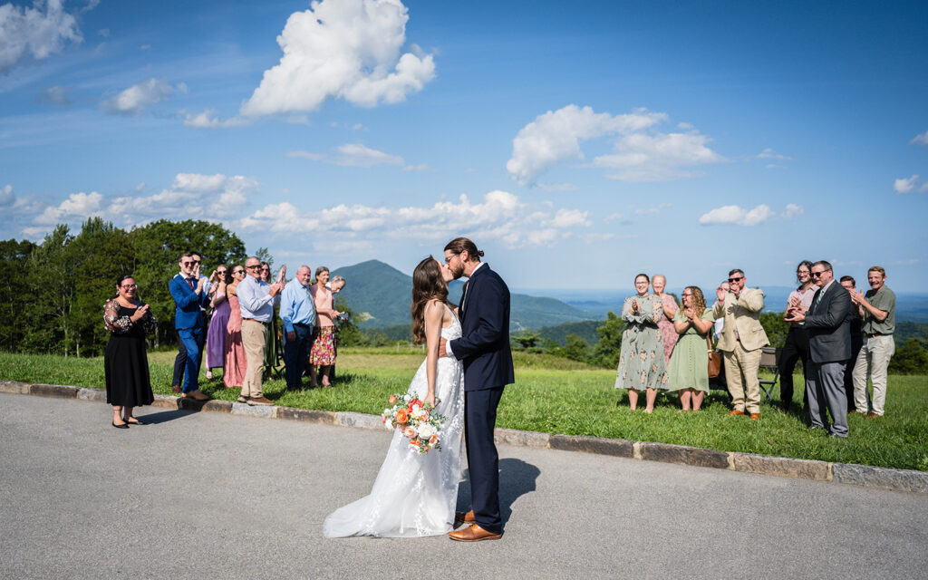 An elopement couple at Cahas Mountain Overlook kiss while their wedding guests look, smiling and clapping.
