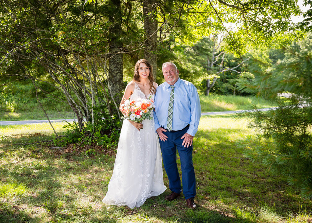 A marrier wearing a wedding dress and holding an artificial wedding bouquet stands with her Dad and smiles for a photo before being walked down the aisle.
