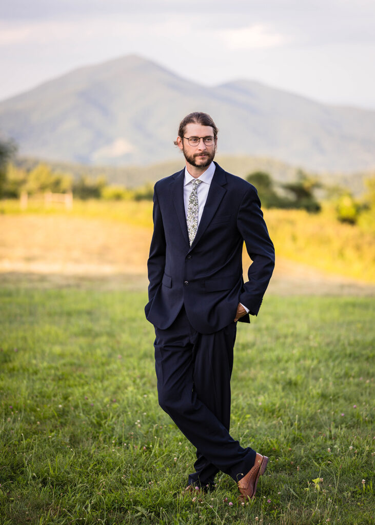 A man wearing a navy blue suit and brown shoes poses in front of Cahas Mountain for golden hour wedding portraits.