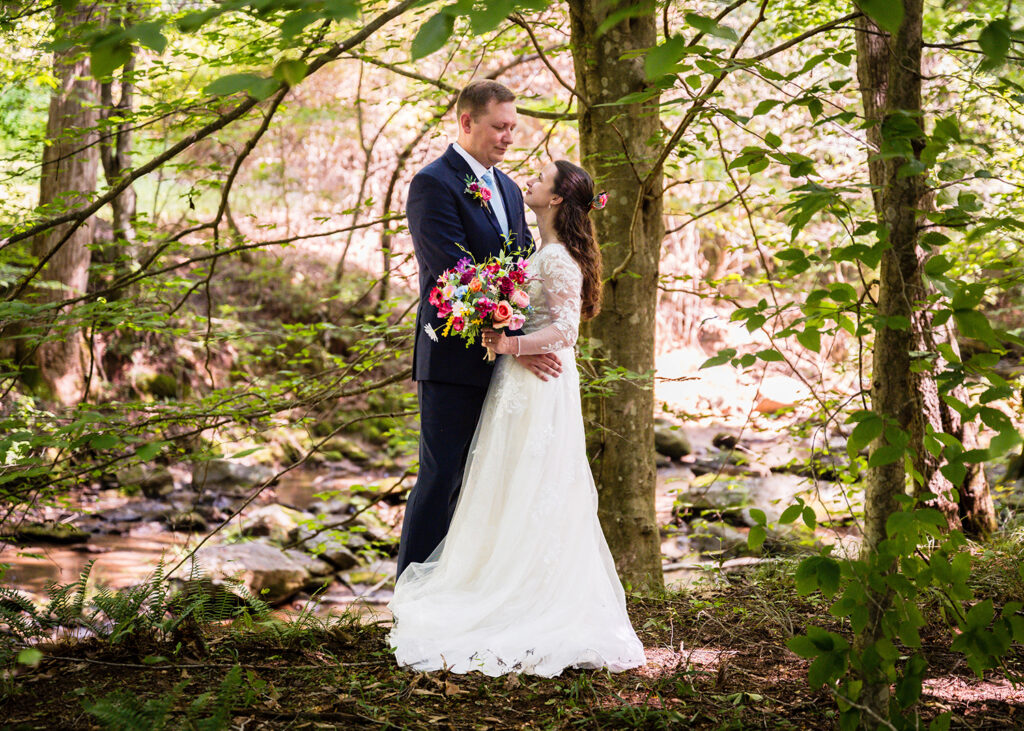 A couple stands in a forest in front of a roaring creek for a romantic photo during their Martinsville, Virginia elopement day.