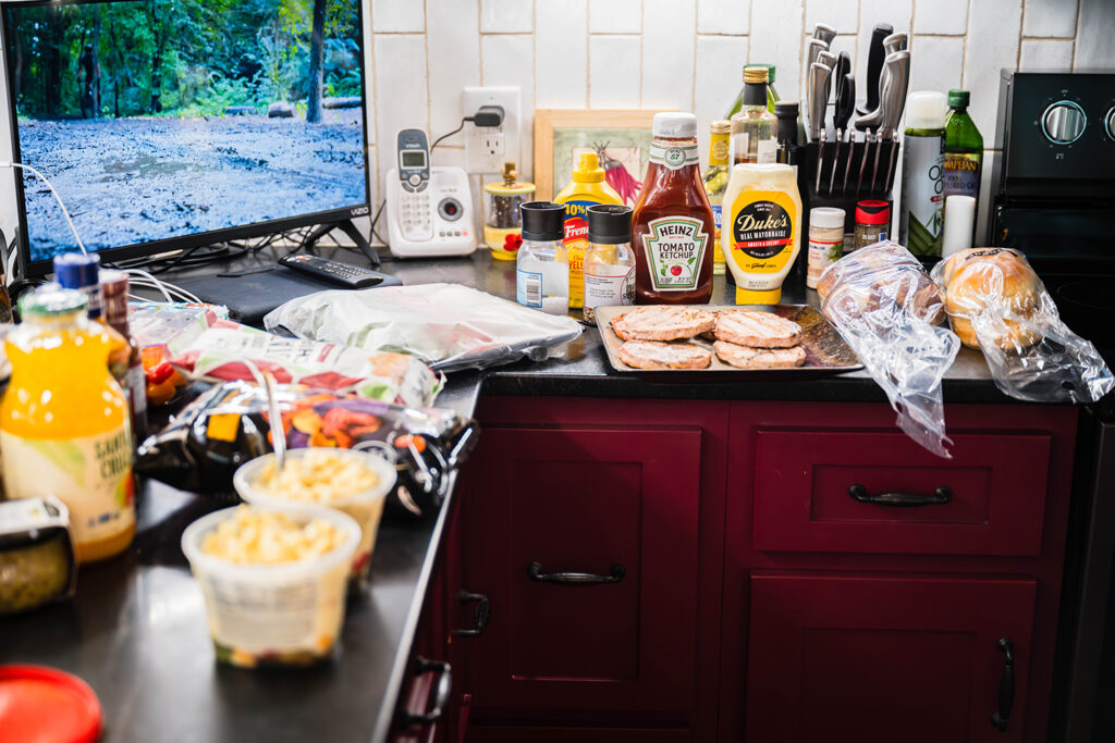 A kitchen counter holding pasta salad, bags of chips, sliced tomato, onion, and lettuce, a plate of turkey burgers, two different types of buns, and a variety of condiments.