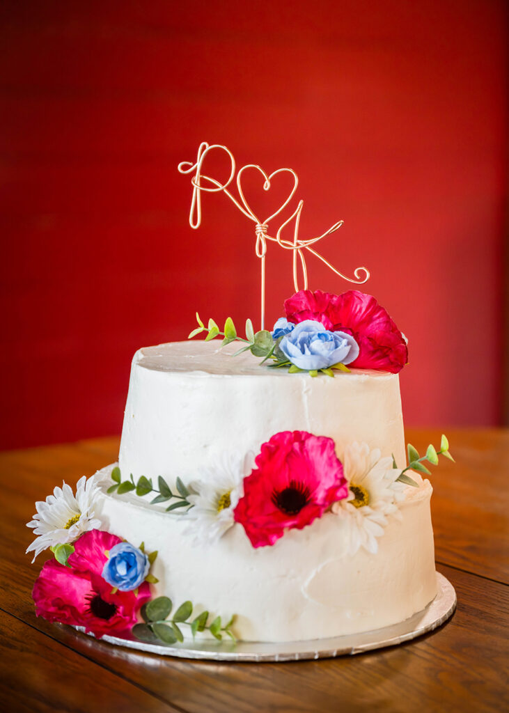 A small two tie vanilla wedding cake with vanilla buttercream icing adorned with artificial flowers sits on a wooden table with a red background. There is a rose gold wire cake topper forming into the shape of a heart connected the letters "R" and "K".