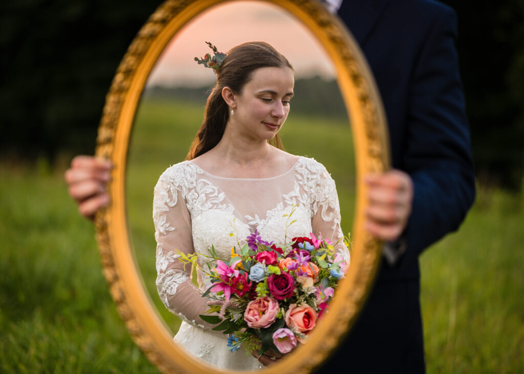 A man holds a vintage mirror which shows a woman looking slightly down and holding her wedding bouquet in the reflection.