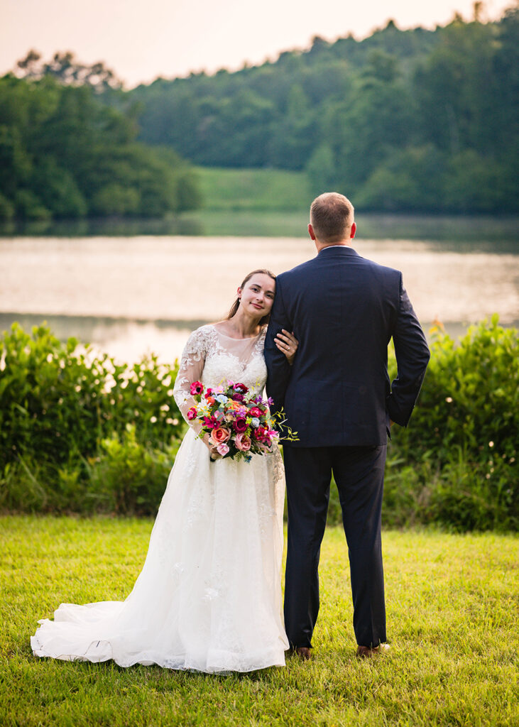 A groom faces the water of a pond as a bride wraps her hand around his arm and lays her head to rest on his shoulder.