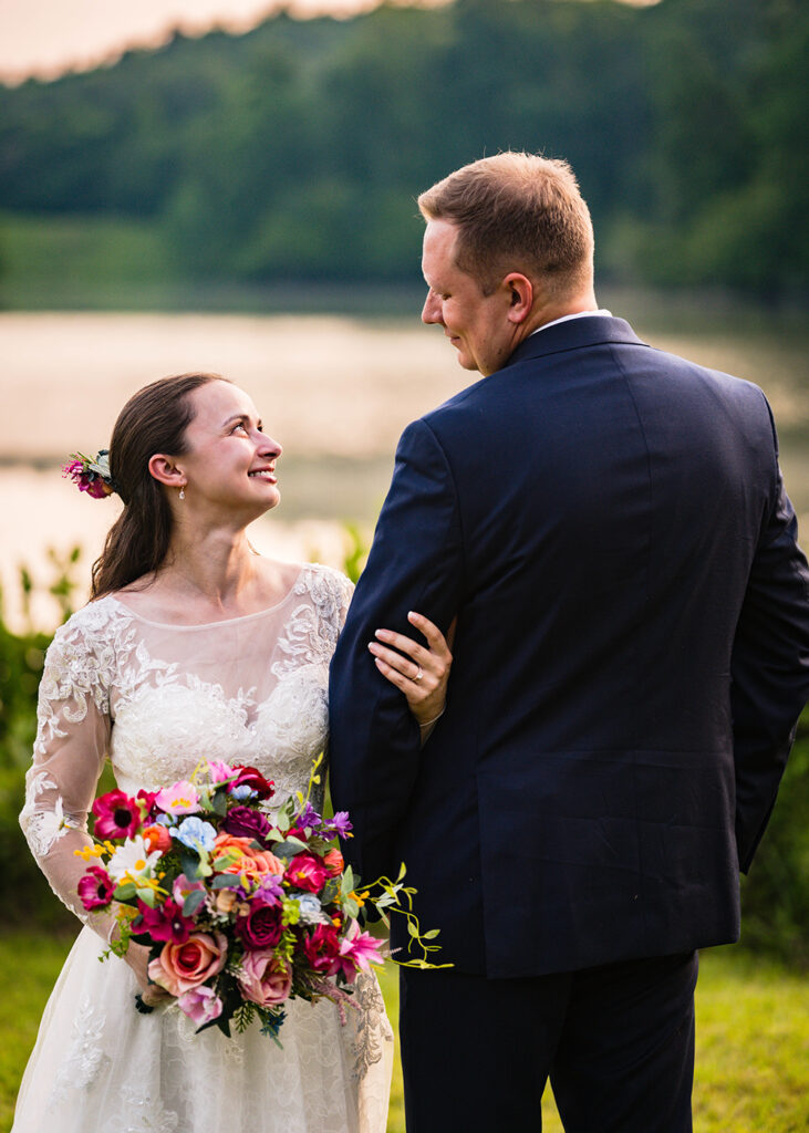 A wedding couple on their Martinsville, Virginia elopement day look at each other and smile in front of a pond at golden hour.