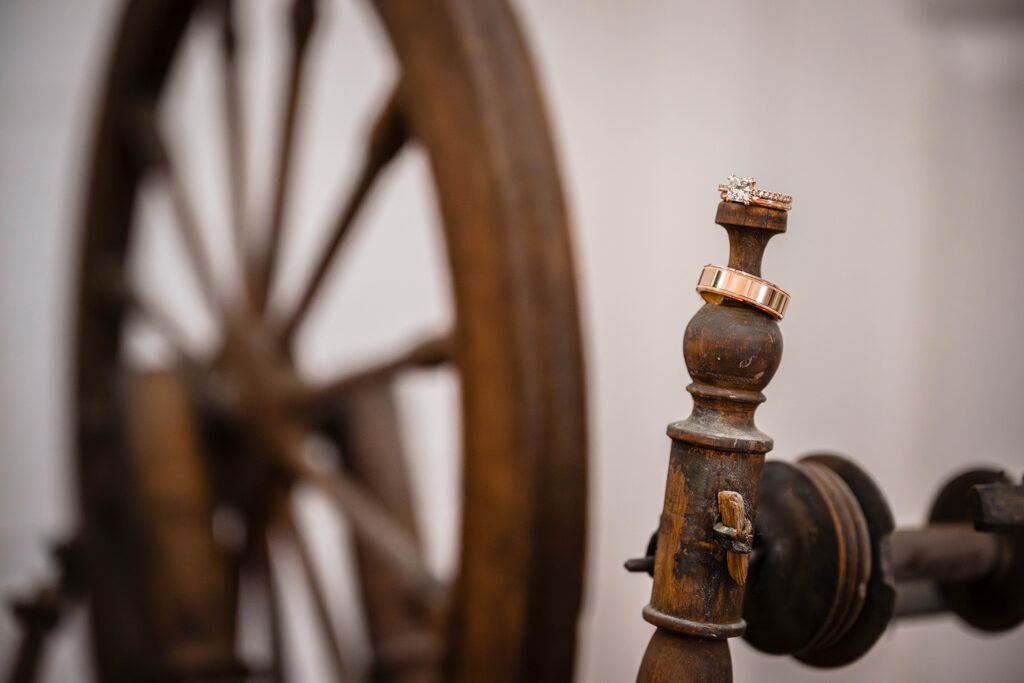 A man's wedding band and a woman's wedding band and engagement ring are placed on a maiden of an antique and vintage spinning wheel.