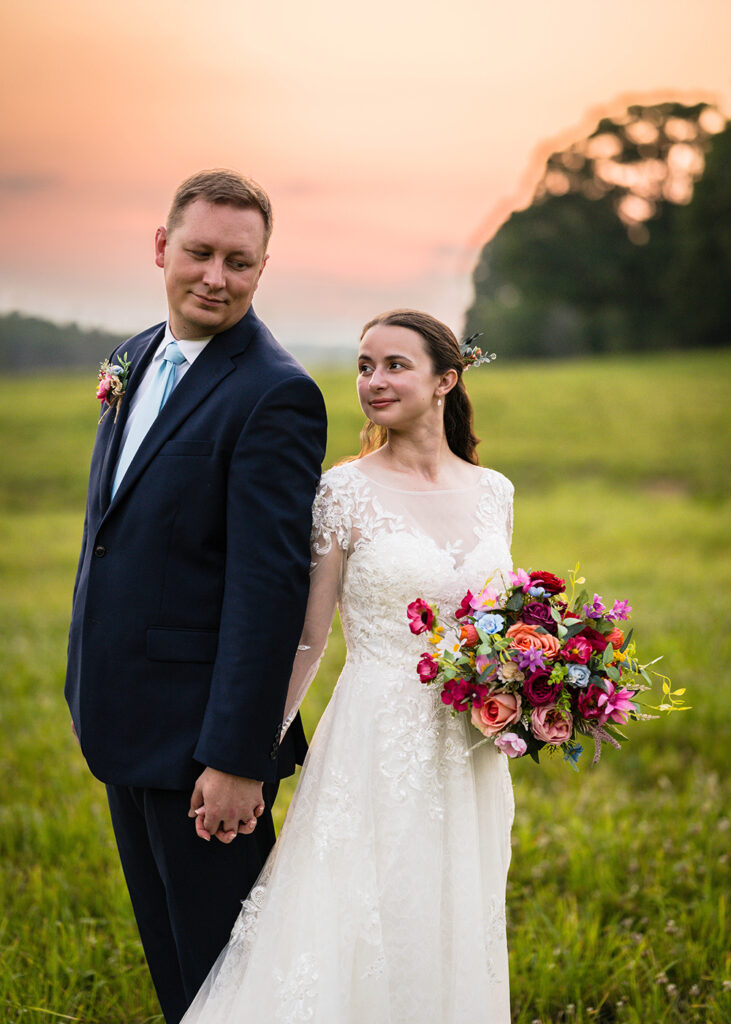 A wedding couple stands back-to-back while holding one another's hands. A man looks down at his partner as she holds her bouquets and looks off in the distance in the opposite direction.