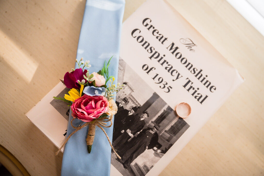 A light blue tie with a boutonniere and a men's wedding band are placed on top of a booked title "The Great Moonshine Conspiracy Trial of 1935" for wedding detail photos.