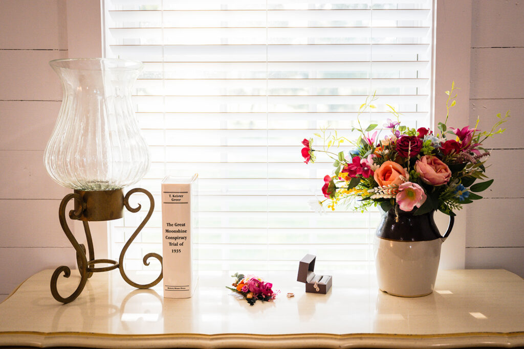 A large lamp, a book titled "The Great Moonshine Conspiracy Trail of 1935", a floral hair clip, rings, earrings in a ring box, and a bouquet of flowers in a vase all placed neatly in a row atop of a dresser in front of an open window.