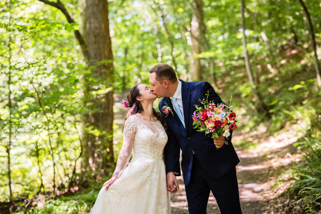 A couple kisses as they hold hands and walk down a forest path during their Martinsville, Virginia elopement day.