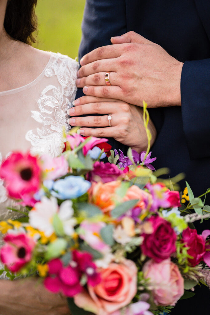 A close up shot of a couple's hands featuring their rose gold wedding rings with a slightly out-of-focus bouquet in the foreground.