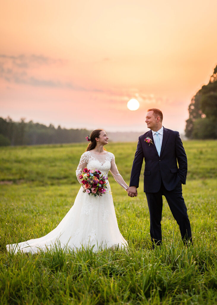 A couple smile widely at one another as they hold each other's hand in front of a golden sunset during their Martinsville, Virginia elopement day.