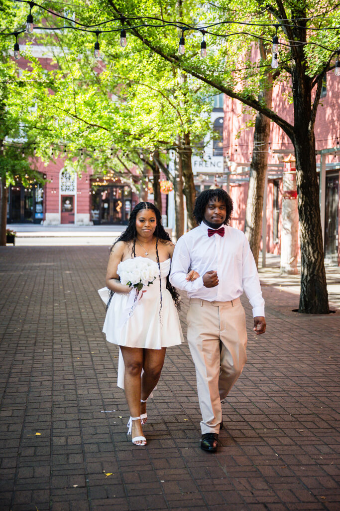 A man holds his arm out for a woman wearing a mini wedding dress and holding a bouquet. The pair walk down the aisle during a Downtown Roanoke wedding at Century Plaza.