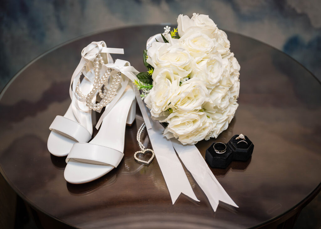A pair of white heels, a faux white flower arrangement, and a ring box with two rings placed inside are placed on a table.