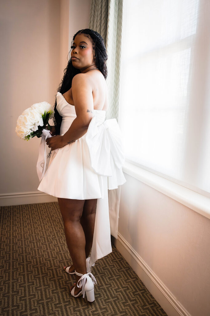 A Black bride poses in a mini wedding dress with a large bow affixed to the back. She is holding a bouquet and wearing white heels.