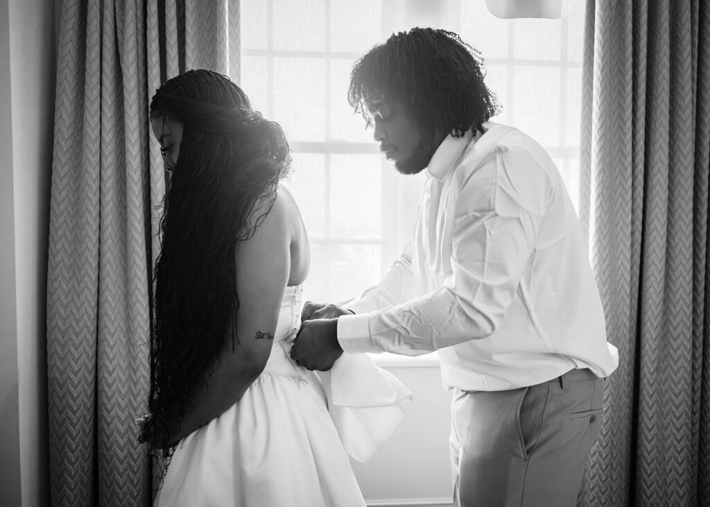 A bride's brother helps her sister affix an attachable, large bow to a dress.