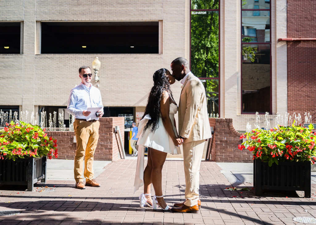 A Black wedding couple go in for their first kiss during their Downtown Roanoke wedding. Their officiant stands to the side and smiles widely.