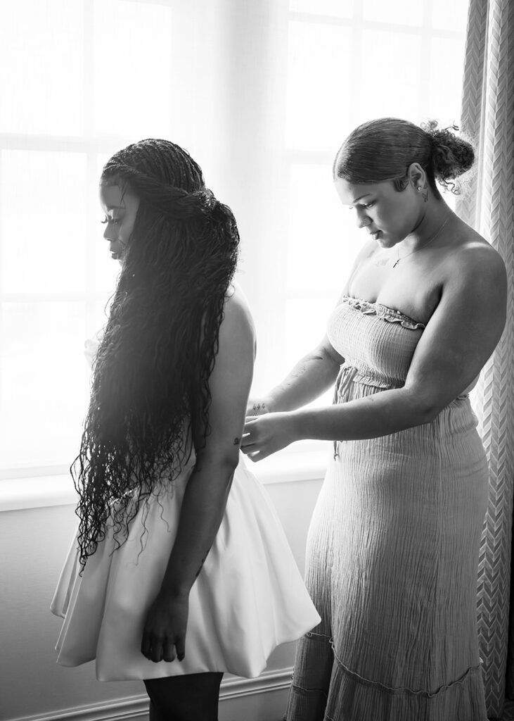 A bride's sister helps her into her wedding dress in a hotel room.