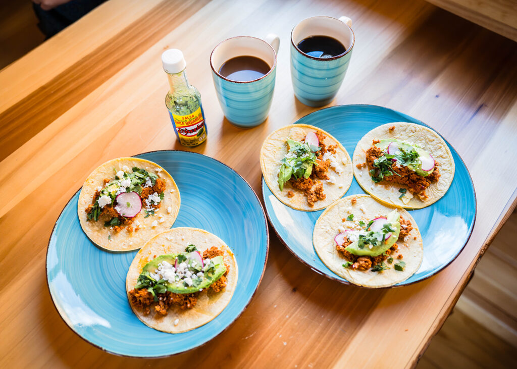 Two plates with breakfast tacos are placed on a table with two cups of coffee and a small bottle of hot sauce.