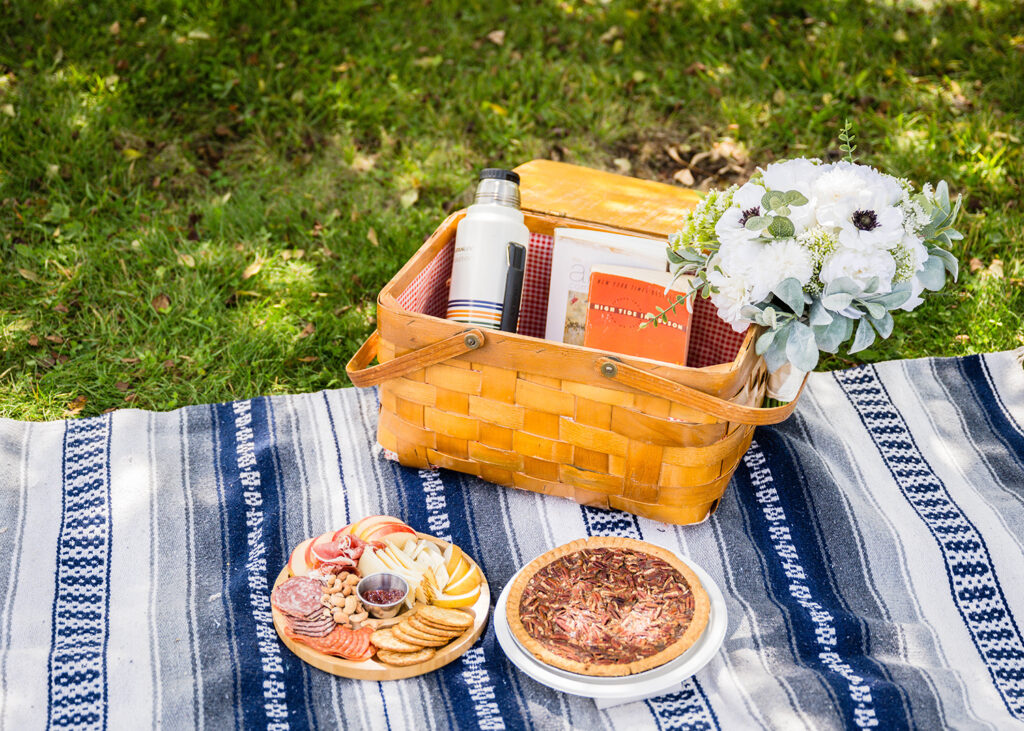 A picnic basket, a charcuterie board full of various meats, nuts, fruits, and crackers, and a full pecan pie sit atop a blanket under the shade of a tree.