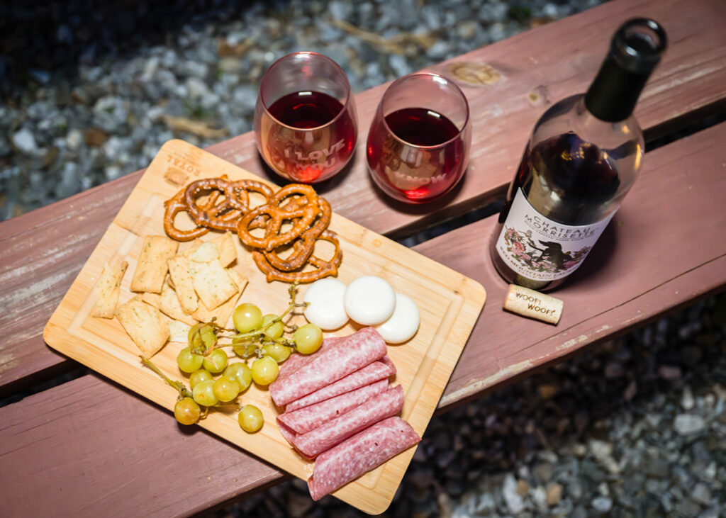 A charcuterie board with meats, cheeses, grapes, crackers, and pretzels sits atop of a red park bench along with two glasses of wine filled with red wine and an open wine bottle. 