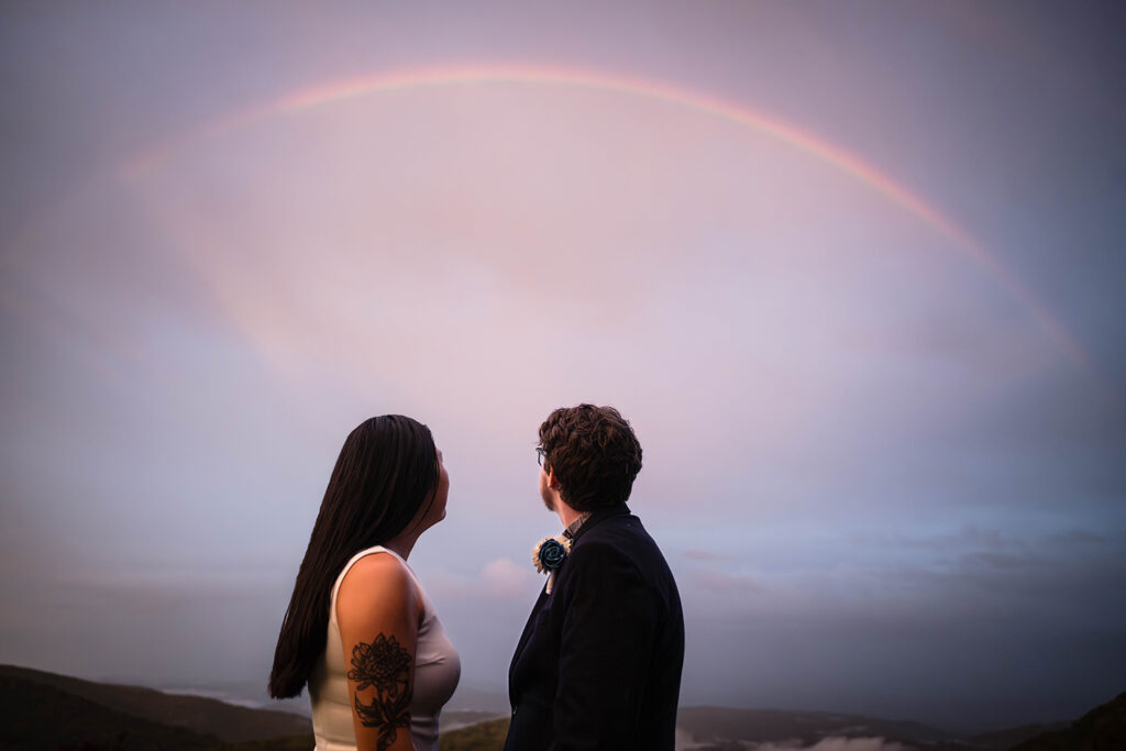 An LGBTQ+ couple looks up towards the sky to see the last bit of a rainbow before it fades away from the sky.