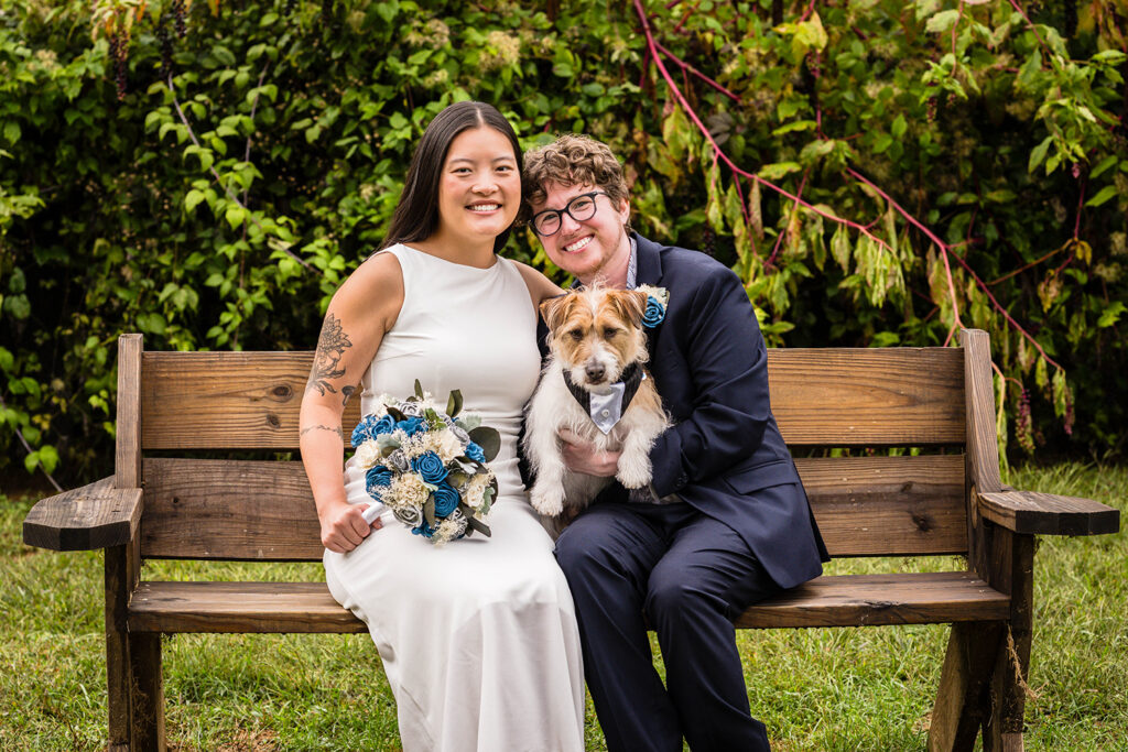 A queer couple sit together on a bench with their dog for a photo.
