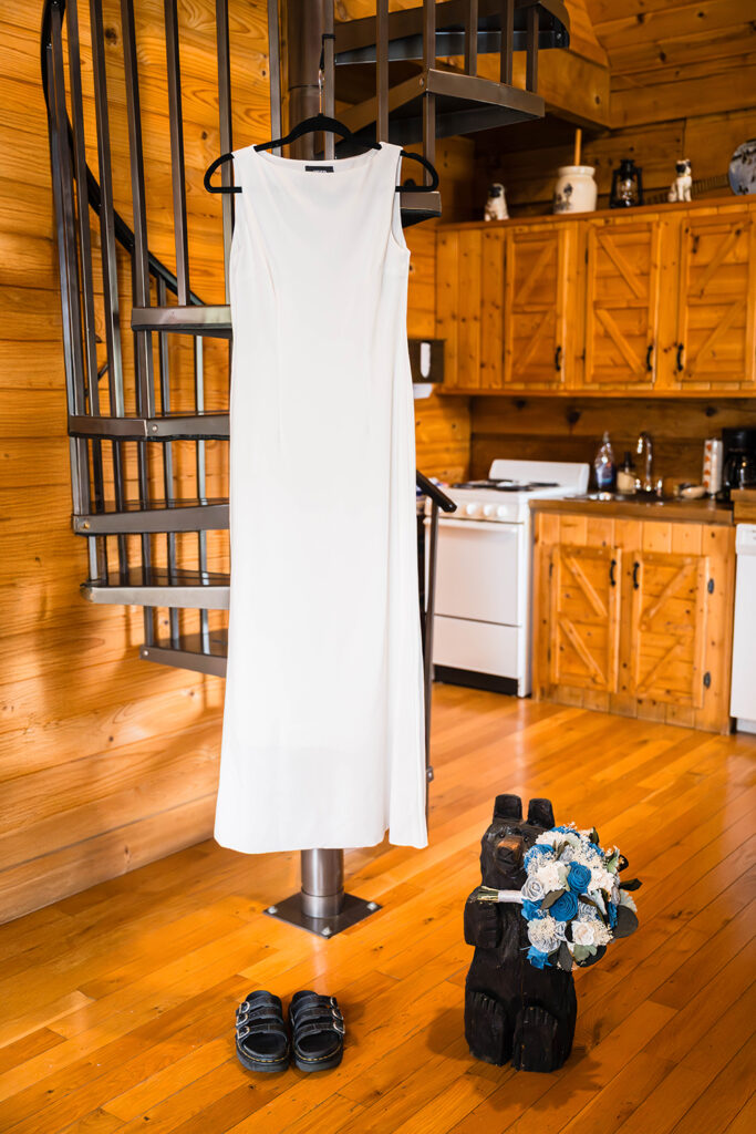 A white wedding dress is hung on a spiral staircase in a HIghland Hideaways airbnb. A pair of black sandals are placed on the floor underneath the dress next to a wood figurine of a black bear holding a wedding bouquet.