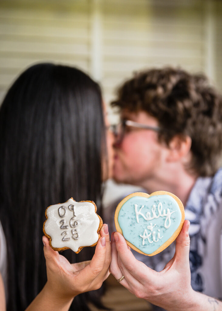 A queer couple goes in for a kiss as they each hold up a cookie. One is a cookie with white icing and the date 09.26.25 written on it with gray royal icing. The other cookie is a heart-shaped cookie with blue frosting with their names "Kelly & Ali" written on the cookie with royal icing.