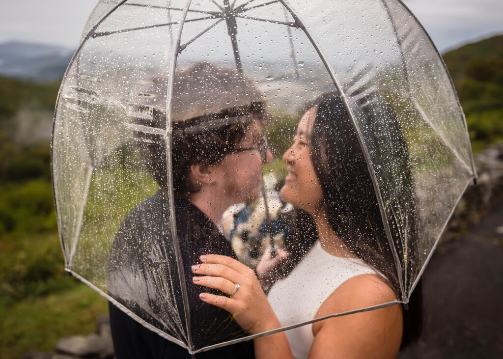 An LGBTQ+ couple look at one another and smile under an umbrella with rain droplets forming at Sugarlands Overlook in Grayson Highlands State Park.