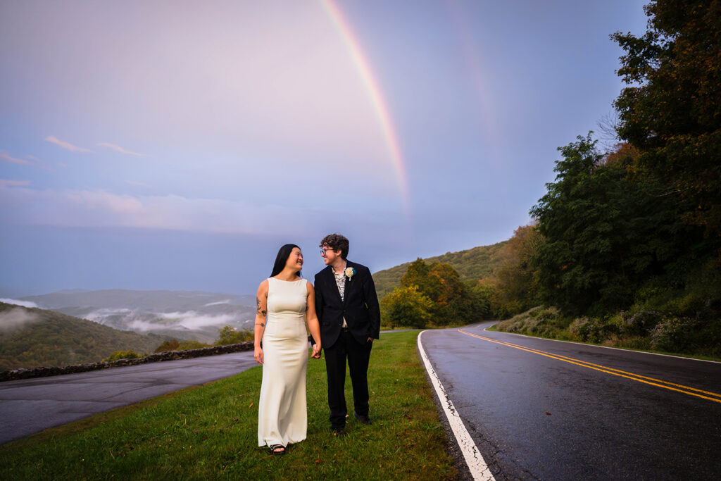 An elopement couple walks together hand-in-hand on the side of the road in Grayson Highlands State park with a double rainbow in the background.