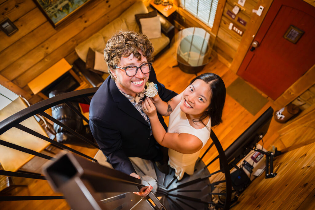 An LGBTQ+ couple poses together on a spiral staircase. The couple looks up and smiles as one of them affixes a boutonniere on their partner.