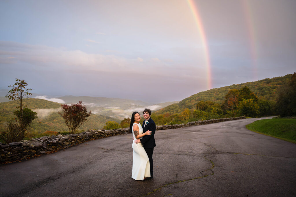 An elopement wedding couple stand together and smile for a photo as two rainbows start to appear in the background.