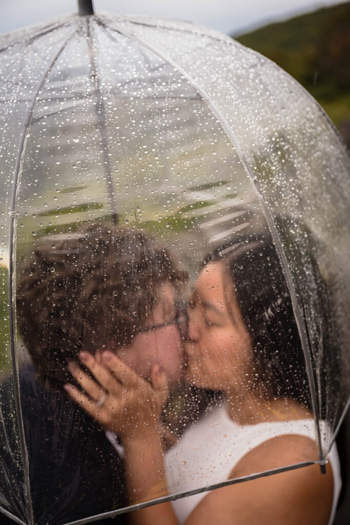 A queer elopement wedding couple kiss one another under a clear umbrella with rain droplets on the outside.
