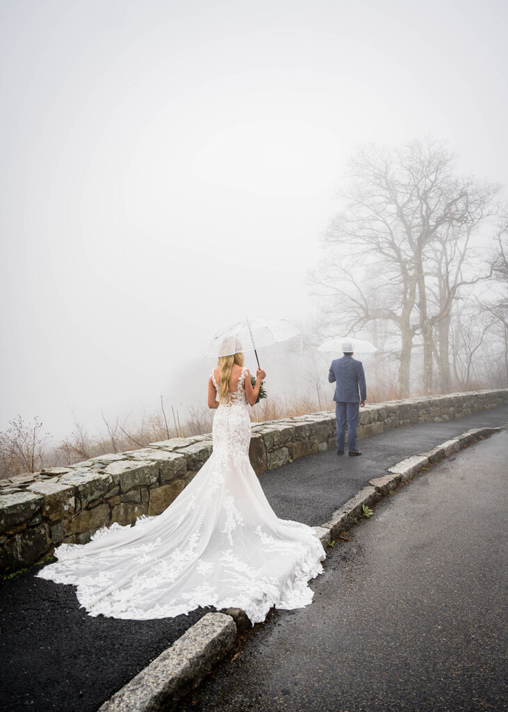 A man wearing a navy blue suit stands facing away from a woman wearing a wedding dress. Both are under clear umbrellas along Thorofare Mountain Overlook.